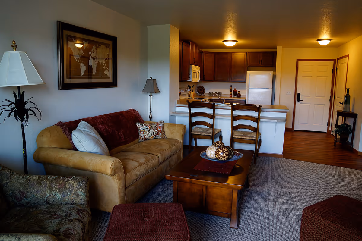 A cozy living room area with a beige couch adorned with pillows and a red throw blanket, a patterned armchair, and a wooden coffee table with decorative spheres. Behind the living room is a kitchen with wooden cabinets, a white refrigerator, a microwave, and a stove. Two wooden chairs are placed at the kitchen counter. The room has warm lighting and a framed world map on the wall.