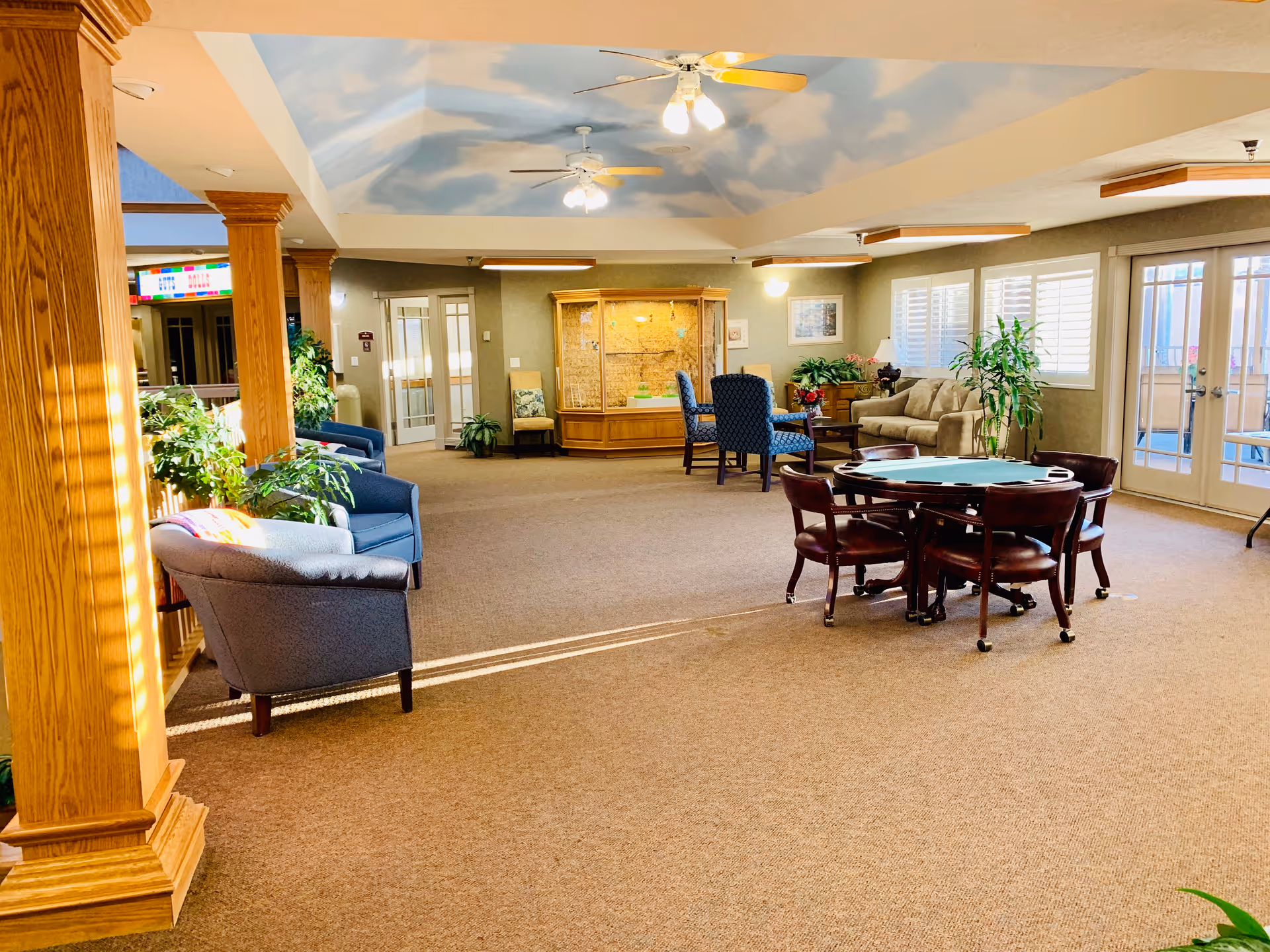 A spacious and well-lit common area in an assisted living community featuring comfortable seating including armchairs and sofas, a round table with chairs, several plants, and large windows with white shutters. The ceiling is painted with a sky and clouds motif and has ceiling fans with lights. Wooden columns and trim add warmth to the space.