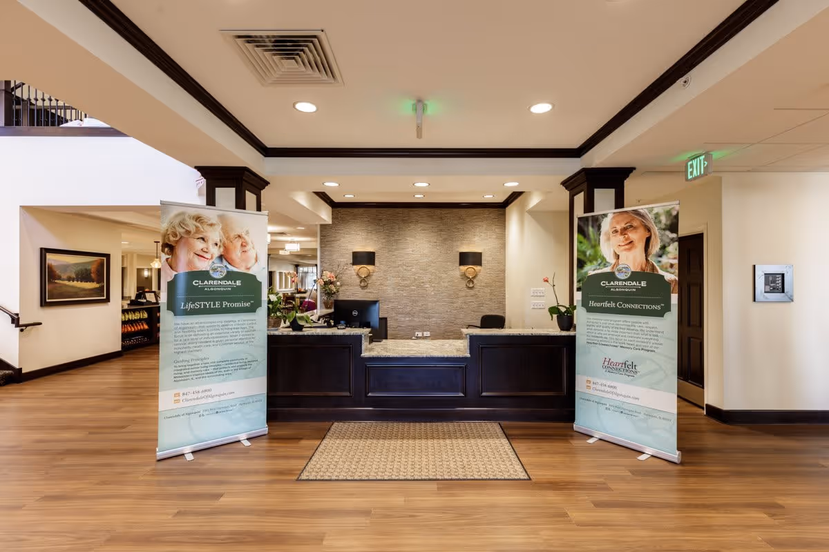 Reception area of Clarendale of Algonquin with a dark wood front desk, two standing banners on either side featuring elderly individuals and information about the facility, wood flooring, and a beige rug in front of the desk. The walls are light-colored with dark trim, and there are wall sconces and ceiling lights illuminating the space.