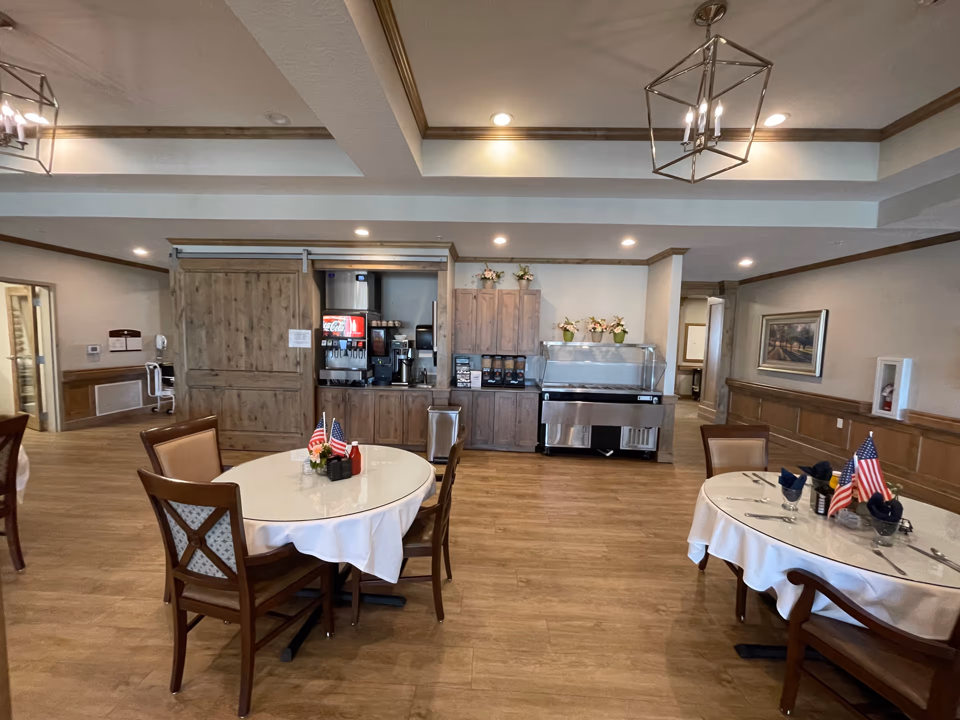 Interior view of a dining area in an assisted living facility with round tables covered with white tablecloths, wooden chairs, and small American flag centerpieces. The background shows a beverage station with coffee machines, a soda dispenser, and a refrigerated display case. The room has wooden flooring, recessed lighting, and decorative ceiling fixtures.
