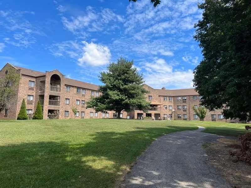 A large three-story brick building with multiple windows and balconies, surrounded by a green lawn and trees under a partly cloudy blue sky. A paved pathway curves through the grass leading towards the building.