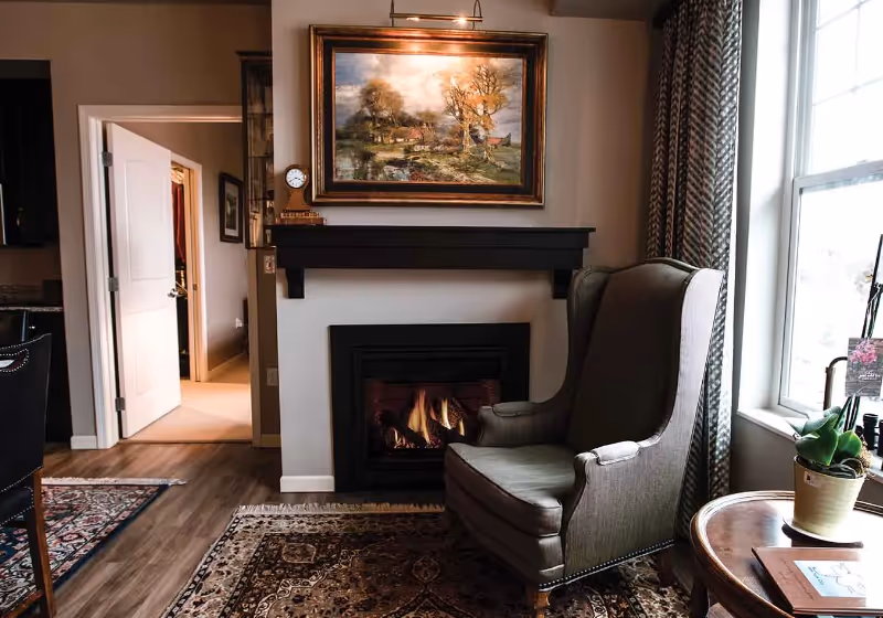 Cozy living room area with a lit fireplace, a comfortable wingback chair, a round wooden side table with a potted plant and books, a large window with patterned curtains, and a framed landscape painting above the fireplace. An open door leads to another room with carpeted flooring.