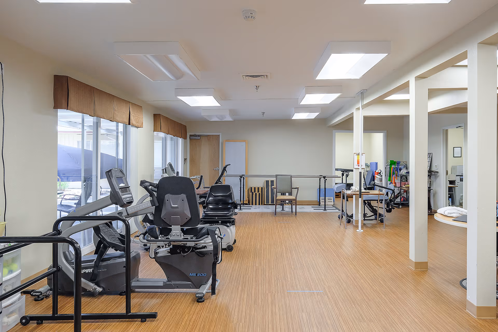 Bright rehabilitation exercise room with cardio machines, parallel bars, chairs, and therapy equipment.