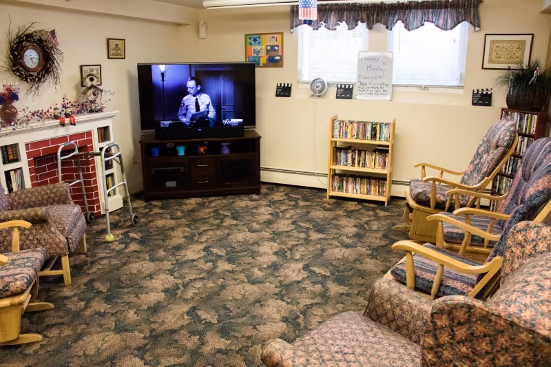 A cozy common room with patterned carpet and several cushioned rocking chairs arranged around the perimeter. A flat-screen TV is on a wooden stand showing a black and white program. There is a small bookshelf filled with books under a window with curtains. A walker is positioned near a decorated fireplace with a wreath and small American flags. Various decorations and framed pictures adorn the walls.