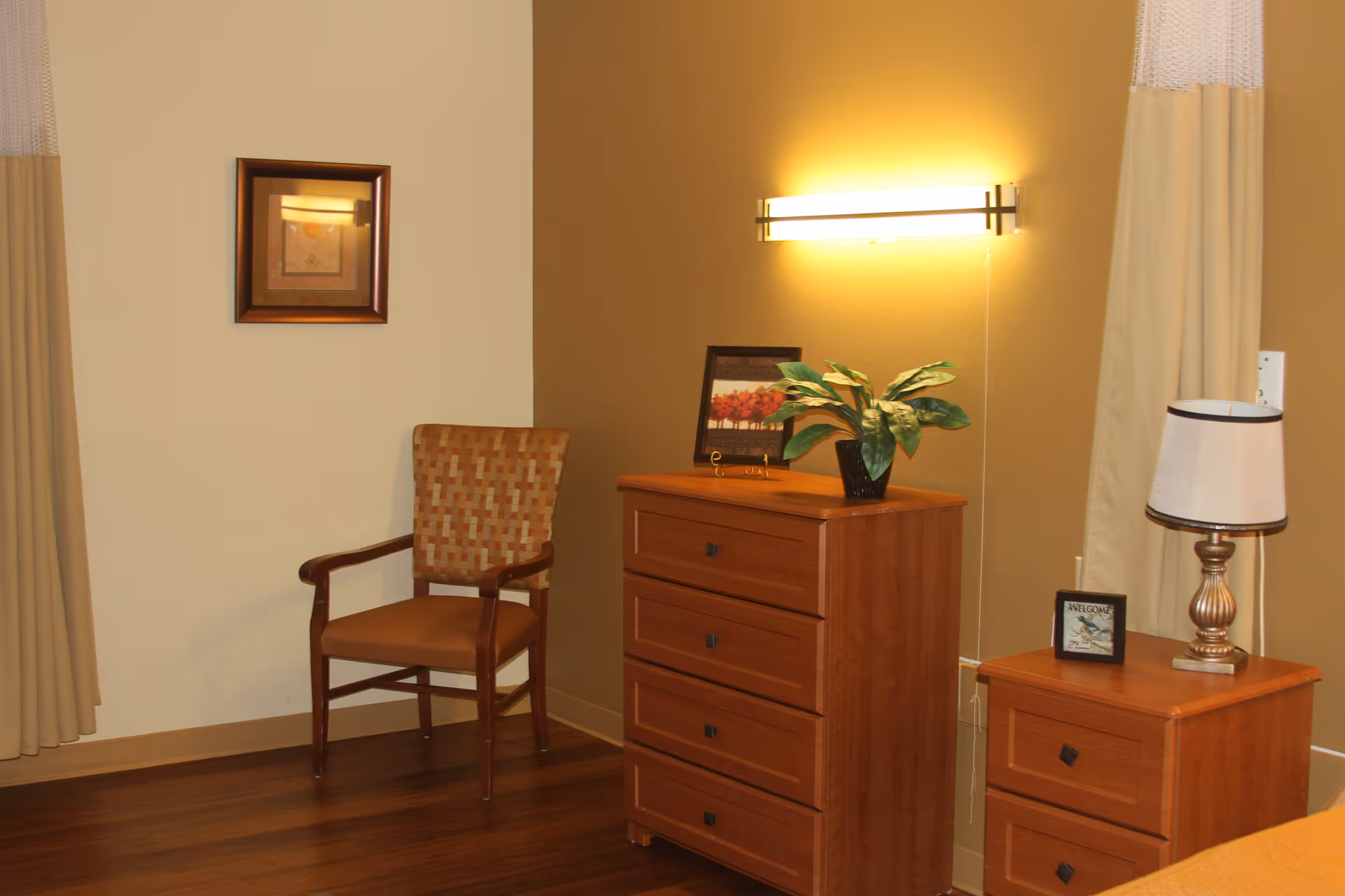A cozy corner of a room featuring a wooden chair with a patterned backrest, a wooden dresser with four drawers topped with a potted plant and a framed picture, and a smaller wooden nightstand with two drawers holding a lamp and a small framed sign. The walls are painted in two tones, beige and light brown, with a wall-mounted light fixture above the dresser and curtains partially visible on the sides.