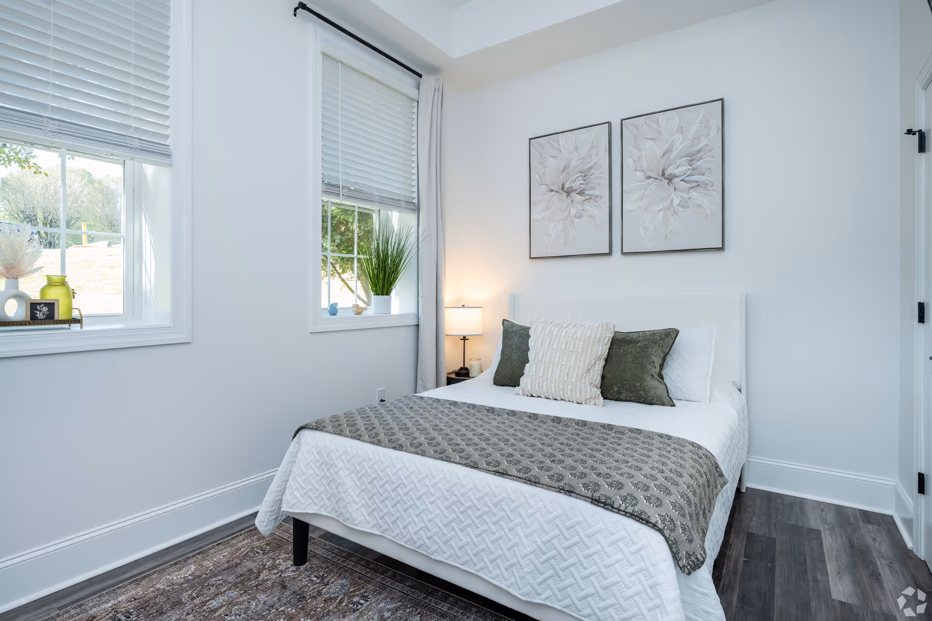 A bright bedroom with a white bed featuring a textured white bedspread, a gray patterned throw blanket, and three decorative pillows. Two framed floral artworks hang above the bed. There are two windows with white blinds and a small nightstand with a lamp beside the bed. The room has light-colored walls and dark wood flooring.