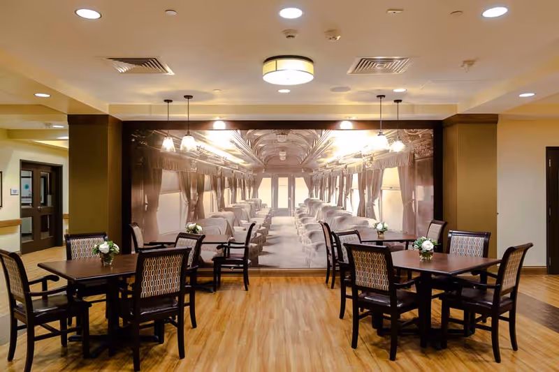 Interior view of a dining area with wooden floors, several dark wood tables and chairs with patterned upholstery, small flower arrangements on each table, and a large sepia-toned mural of a vintage train car seating area on the far wall.