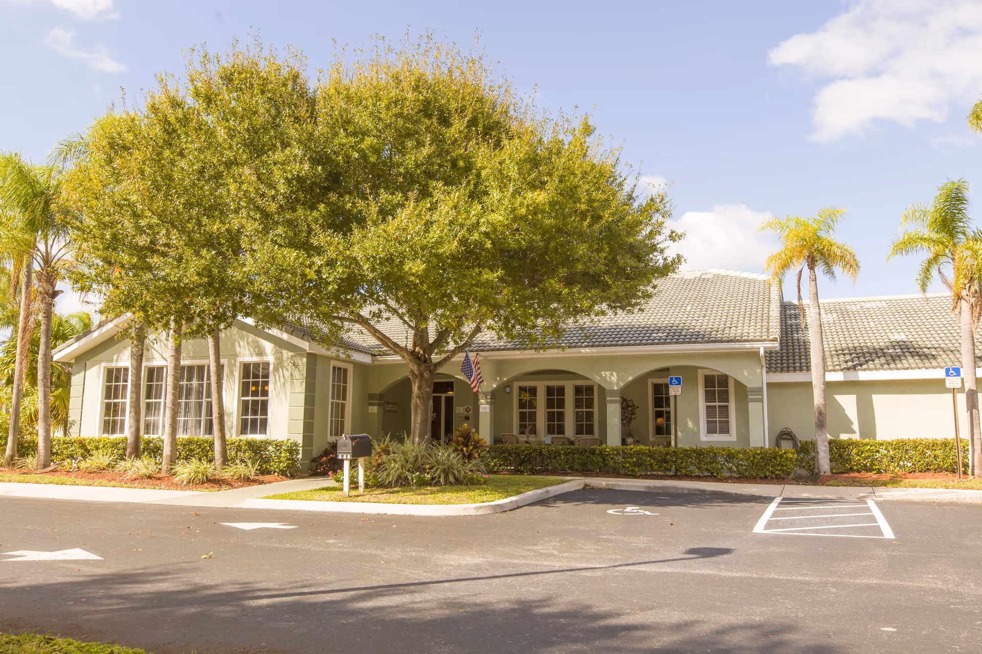 Front exterior of a single-story senior living facility with a tiled roof, large trees and palm trees, and a parking area with accessible spaces.