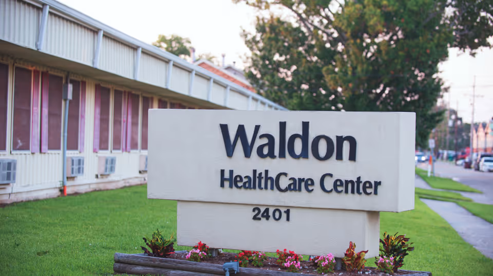 Outdoor view of a sign for Waldon HealthCare Center with the address number 2401, situated on a grassy area with flowers and a building with windows and air conditioning units in the background.