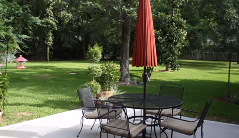 Outdoor patio area with a round metal table and four cushioned chairs. A closed red umbrella is positioned in the center of the table. The patio overlooks a green grassy yard with trees and bushes in the background.