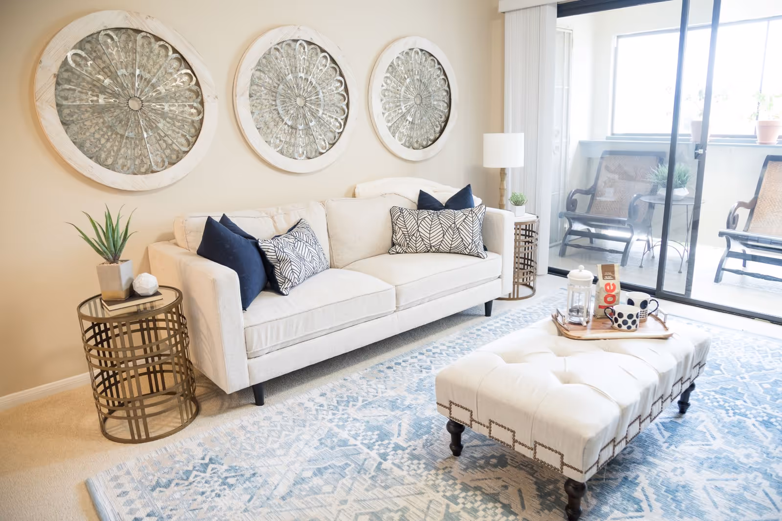Bright living room with a beige sofa, patterned pillows, three round wall artworks, a tufted ottoman, side tables, and sliding glass doors to a balcony.