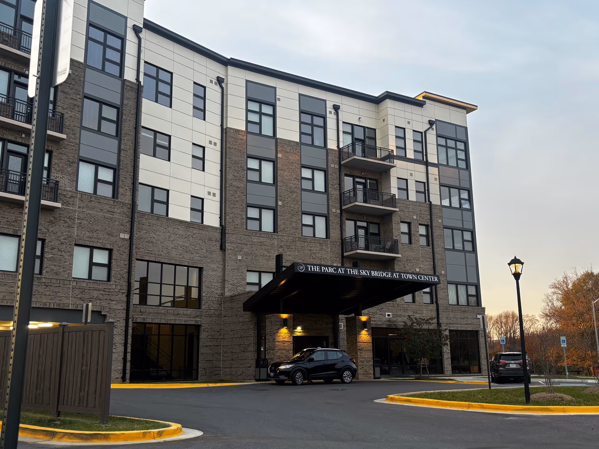 Five-story modern brick-and-panel building front with a covered entrance and parked cars at the Sky Bridge at Town Center.