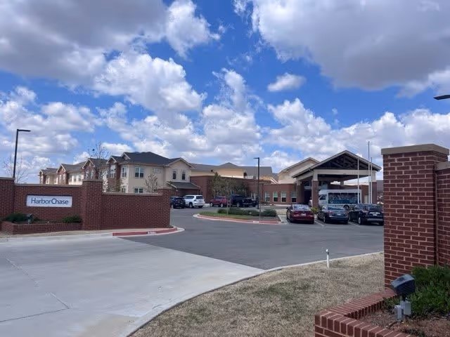 Exterior view of a senior living facility with a brick entrance sign that reads 'HarborChase'. The building has multiple stories with beige and brick walls, several parked cars, and a covered entrance. The sky is partly cloudy.