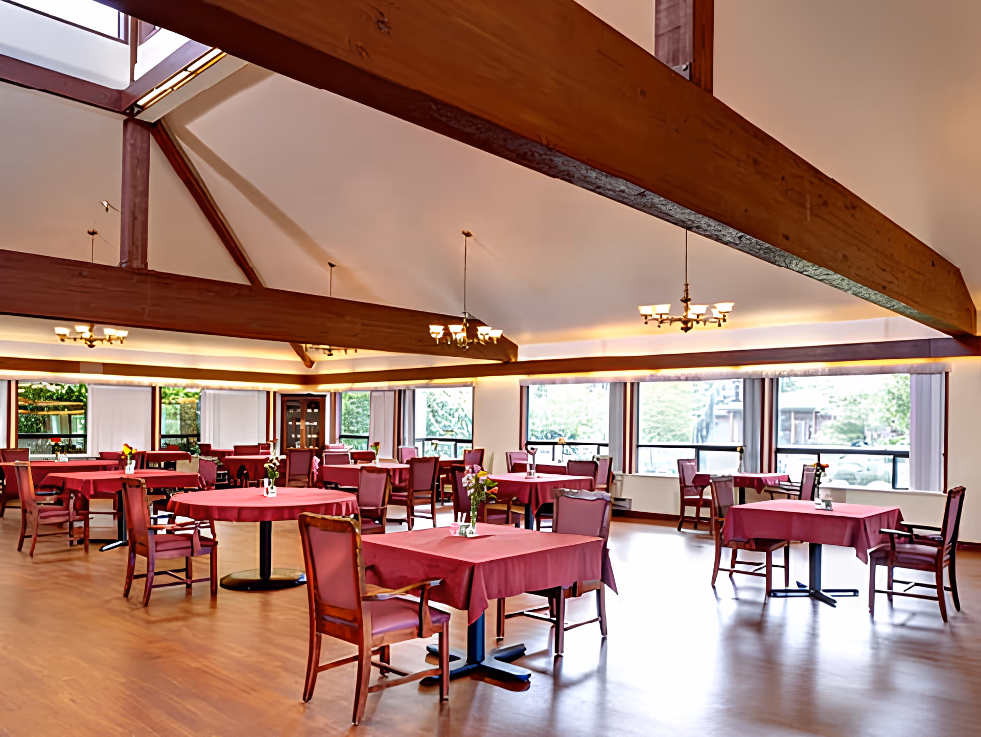 Dining room with red-tablecloth tables and wooden chairs under exposed ceiling beams and large windows.