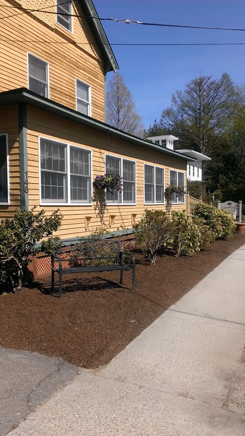 Exterior view of a yellow building with multiple windows and hanging flower pots. There is a black metal bench in front of the building surrounded by mulch and bushes. A sidewalk runs alongside the building under a clear blue sky.