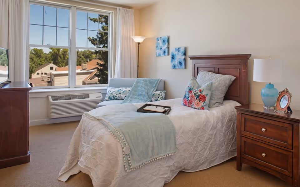 Sunlit furnished bedroom with a single bed, wooden nightstand, armchair by a large window, and decorative pillows and lamp.