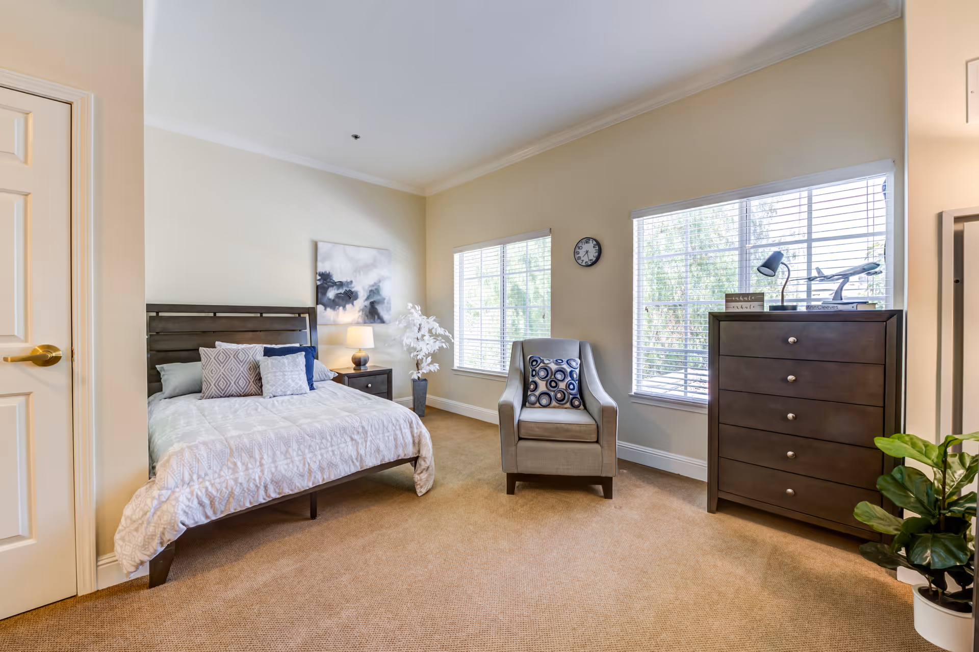 A bright and neatly arranged bedroom with a bed featuring multiple pillows, a nightstand with a lamp, a gray armchair with a decorative pillow, a dark wooden dresser with a lamp and books on top, two large windows with blinds, a wall clock, and a potted plant on the floor.