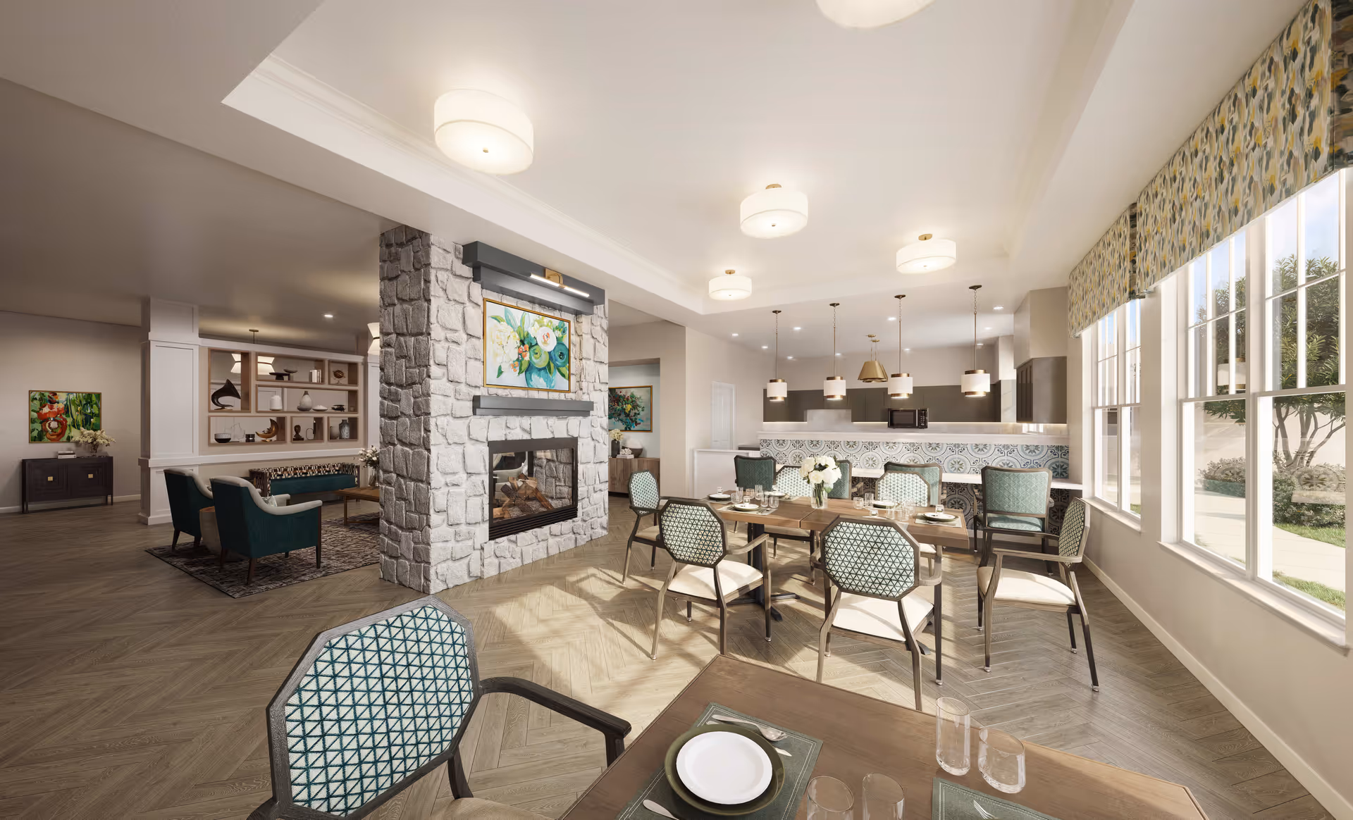 A bright and spacious common area in an assisted living facility featuring a dining area with tables and chairs set for meals, a stone fireplace with a colorful painting above it, and a kitchen area with pendant lights and a patterned tile backsplash. Large windows on the right side let in natural light and offer a view of greenery outside.