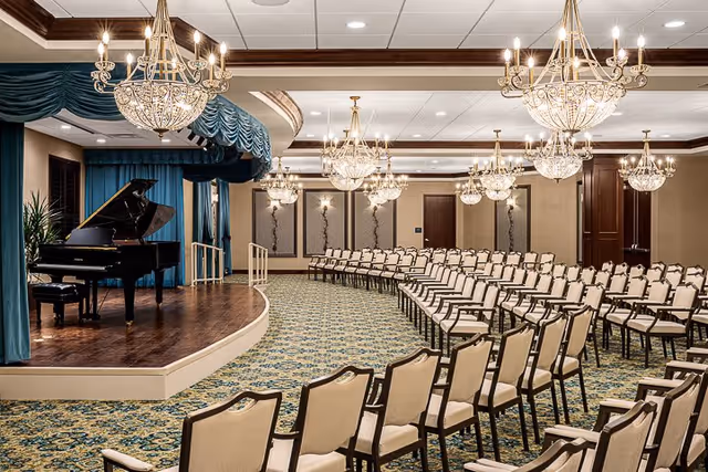A large elegant event room with rows of beige cushioned chairs arranged facing a small stage with a grand piano. The room features ornate chandeliers hanging from the ceiling, patterned carpet, and decorative wall panels.