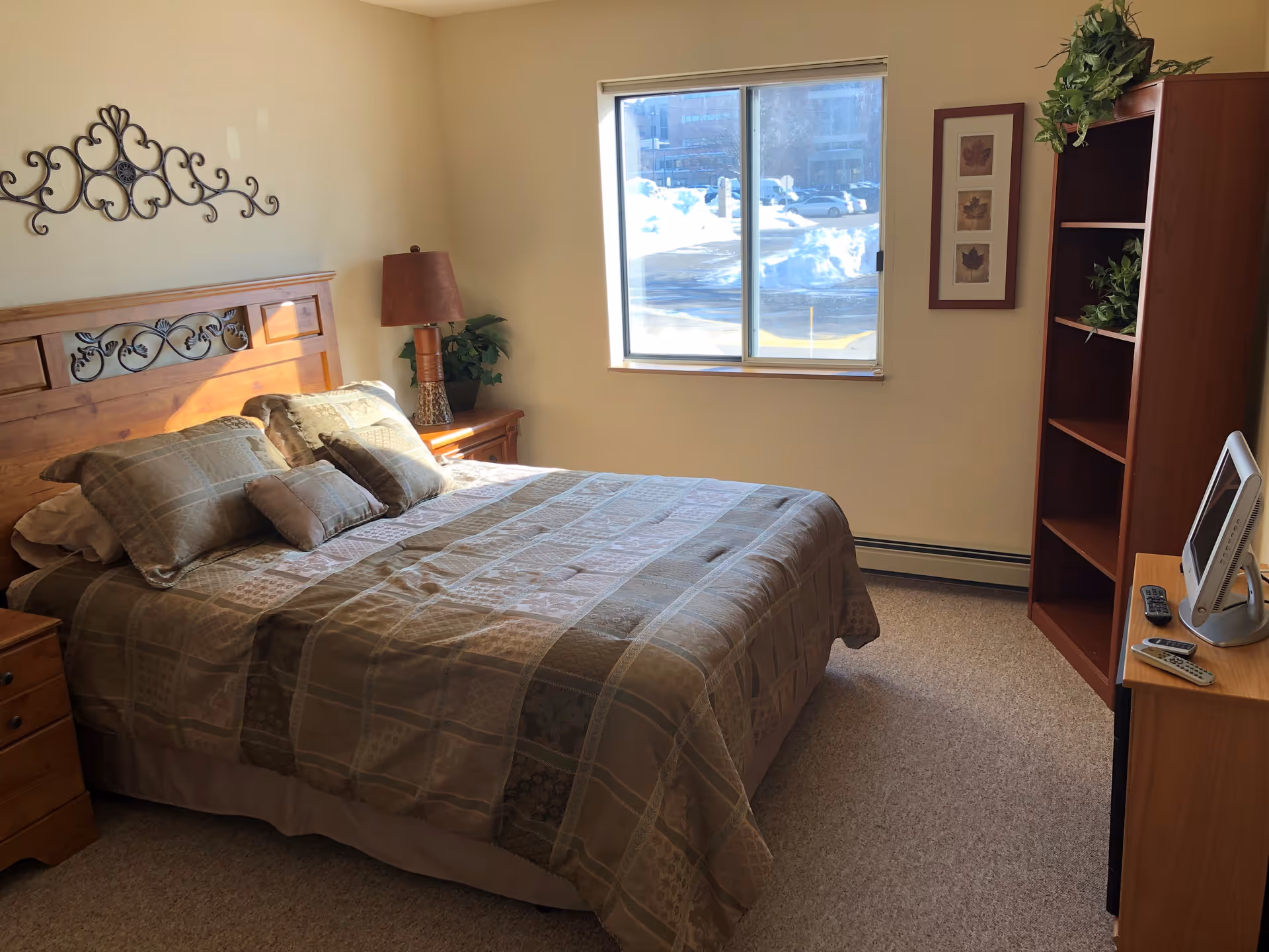 Sunlit bedroom with a made bed, wooden headboard, nightstand, bookshelf and a window showing snow outside.