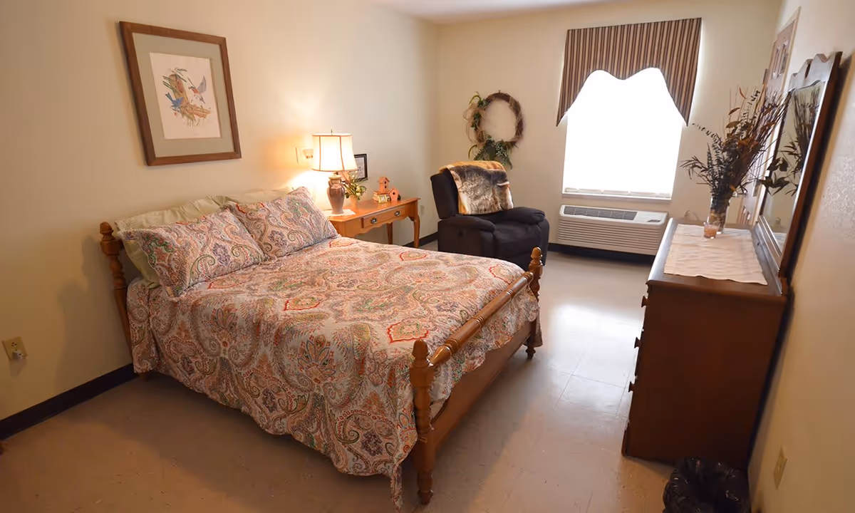 A cozy bedroom in a retirement center featuring a wooden bed with a colorful patterned bedspread and pillows. Next to the bed is a wooden nightstand with a lamp, small plants, and decorative items. There is a comfortable recliner chair with a throw blanket near a window with a valance. A wooden dresser with a mirror and a vase with dried flowers is positioned against the opposite wall. The room has light-colored walls and flooring.
