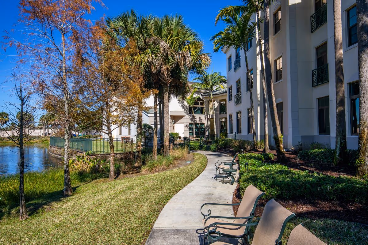 Outdoor pathway with benches alongside a senior living facility building, surrounded by palm trees, other greenery, and a pond under a clear blue sky.