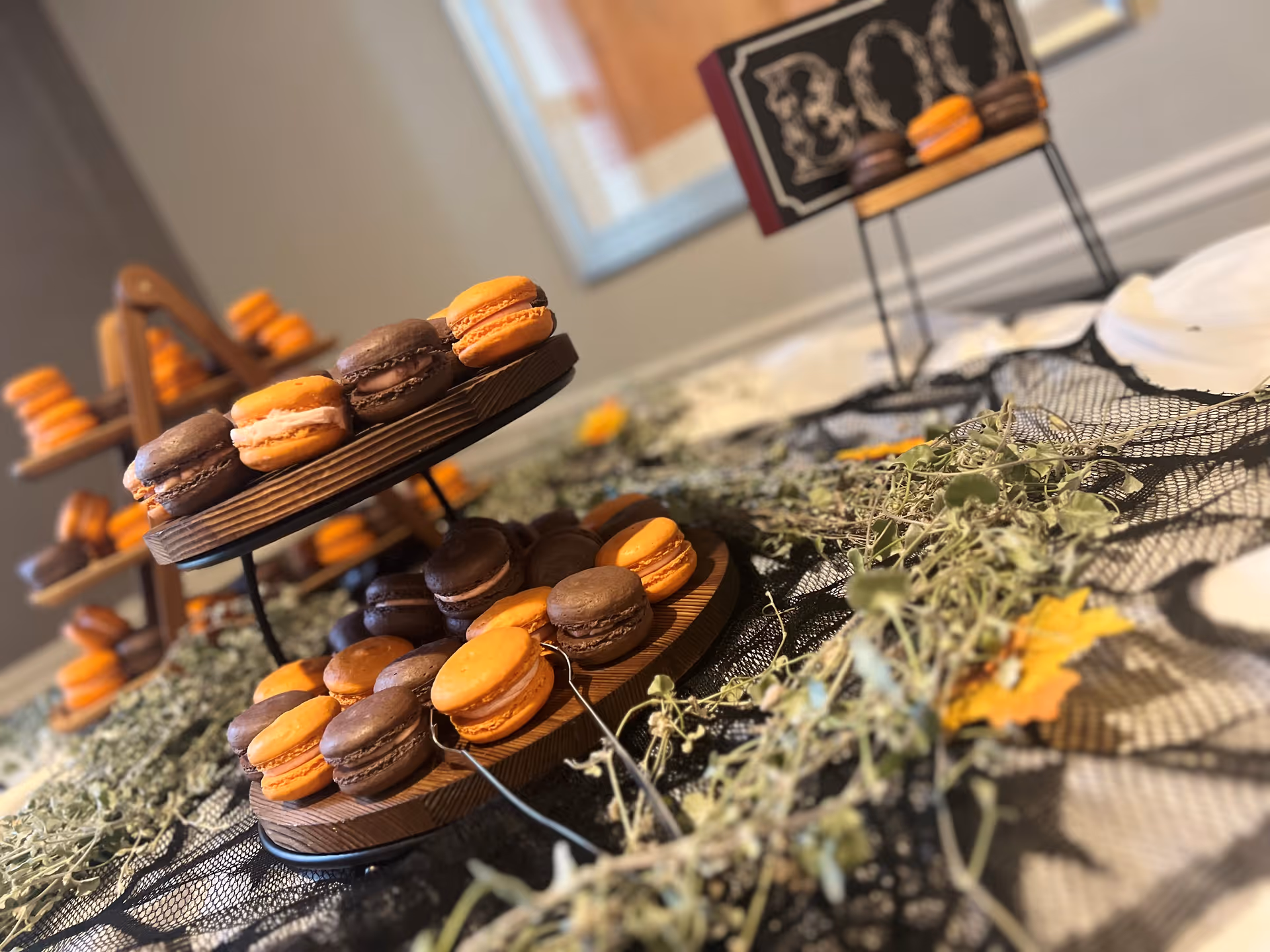 Close-up view of two-tiered wooden stands displaying orange and chocolate macarons on a table decorated with dried leaves and greenery, with additional macaron stands visible in the background in a softly lit room.
