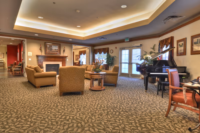 A spacious and warmly lit living room area with patterned carpet, multiple upholstered chairs and sofas arranged around a wooden coffee table, a fireplace with a painting above it, large windows with curtains, and a black grand piano with a floral arrangement on top near the entrance doors.
