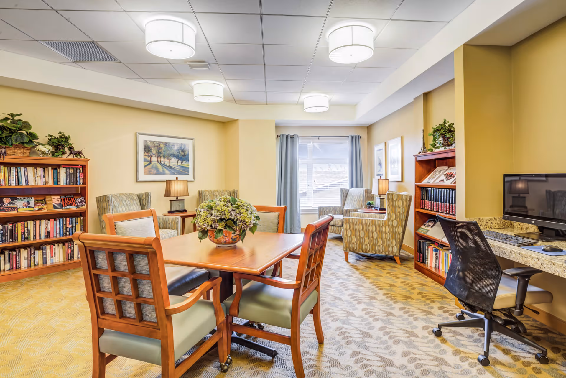 A cozy senior living common area with a wooden table and four chairs in the center, a floral centerpiece on the table, bookshelves filled with books on the left and right sides, two armchairs near a window with curtains, a desk with a computer and office chair on the right, and soft yellow walls with framed artwork.
