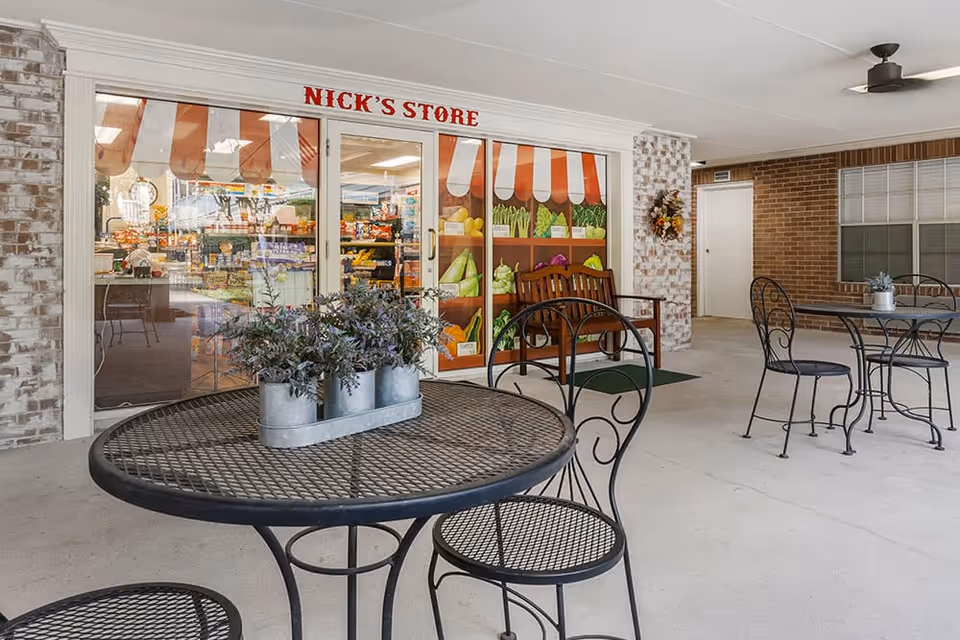Outdoor covered patio area with black metal tables and chairs. A small potted plant centerpiece is on the closest table. In the background, there is a storefront with glass doors labeled 'Nick's Store' and decorated with a painted awning and images of vegetables. The area has brick walls and a ceiling fan.