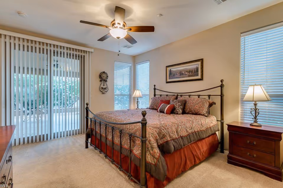 A bedroom with a large bed featuring a patterned bedspread and multiple pillows. The room has beige walls, two windows with blinds, a ceiling fan with a light, two wooden nightstands with lamps, and vertical blinds covering a sliding glass door that leads outside.
