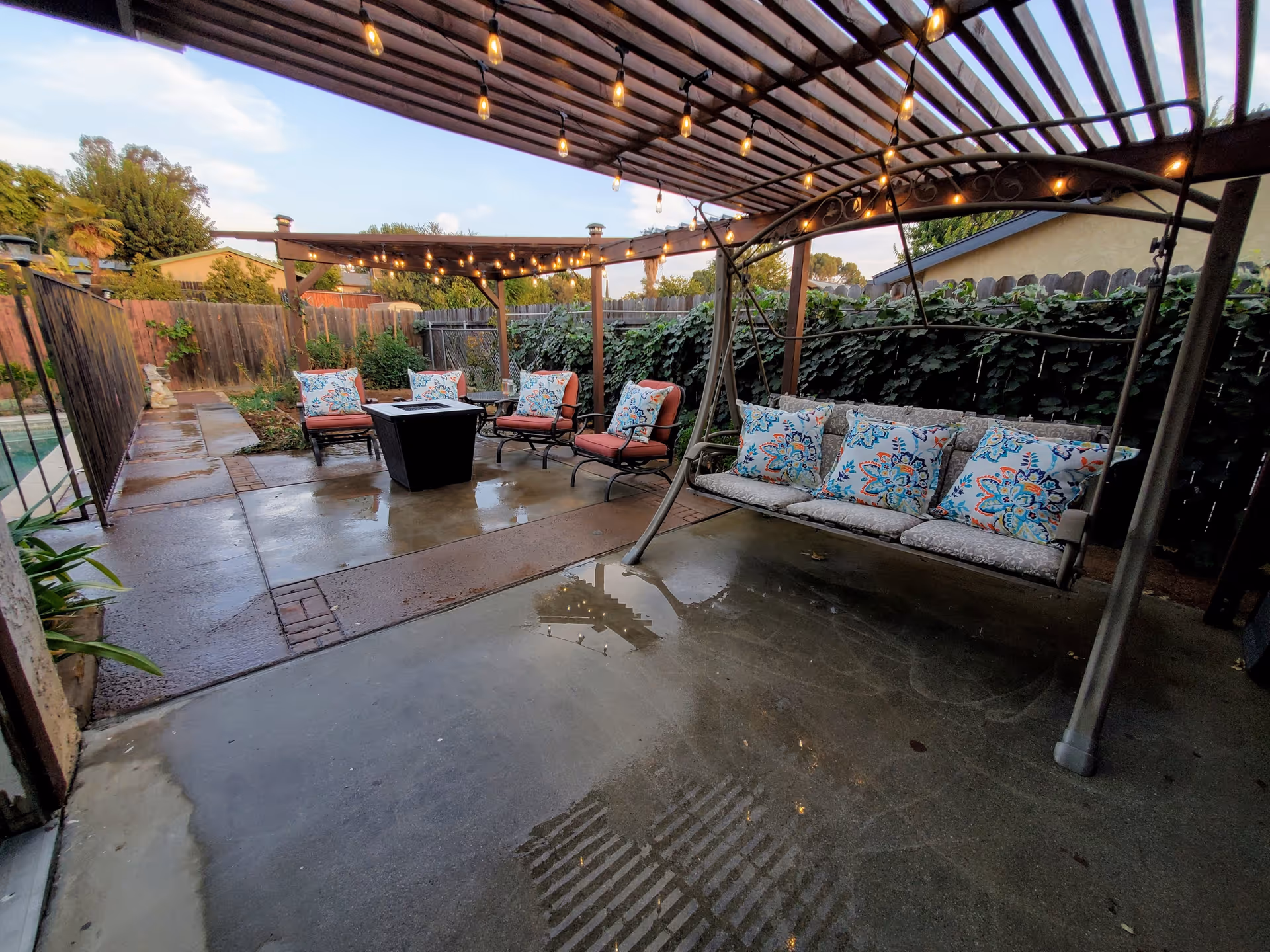 Outdoor patio area with a pergola covered in string lights, featuring cushioned chairs arranged around a fire pit table and a cushioned swing with patterned pillows. The patio is surrounded by a wooden fence and greenery.