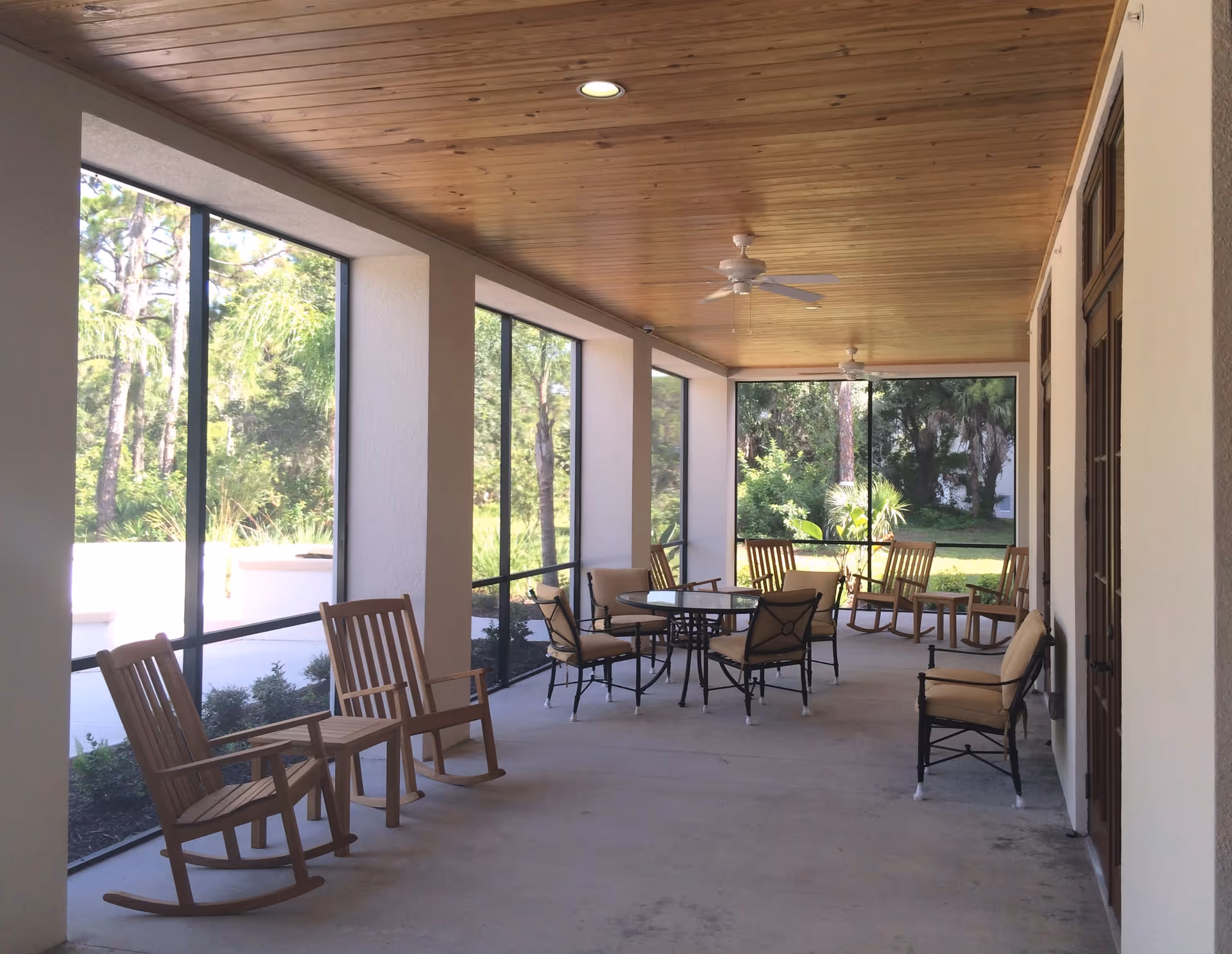 A covered outdoor patio area with a wooden ceiling, ceiling fans, and large screened windows overlooking greenery. The patio is furnished with wooden rocking chairs, cushioned armchairs, and a round table with chairs arranged around it.