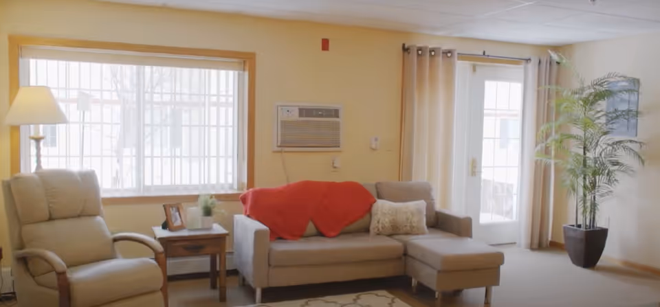 A cozy living room with a beige recliner chair, a small wooden side table with a framed photo and a plant, a gray sectional sofa with a red throw blanket and a white pillow, a floor lamp, a large window with blinds, a glass door with curtains, and a tall potted plant in the corner.