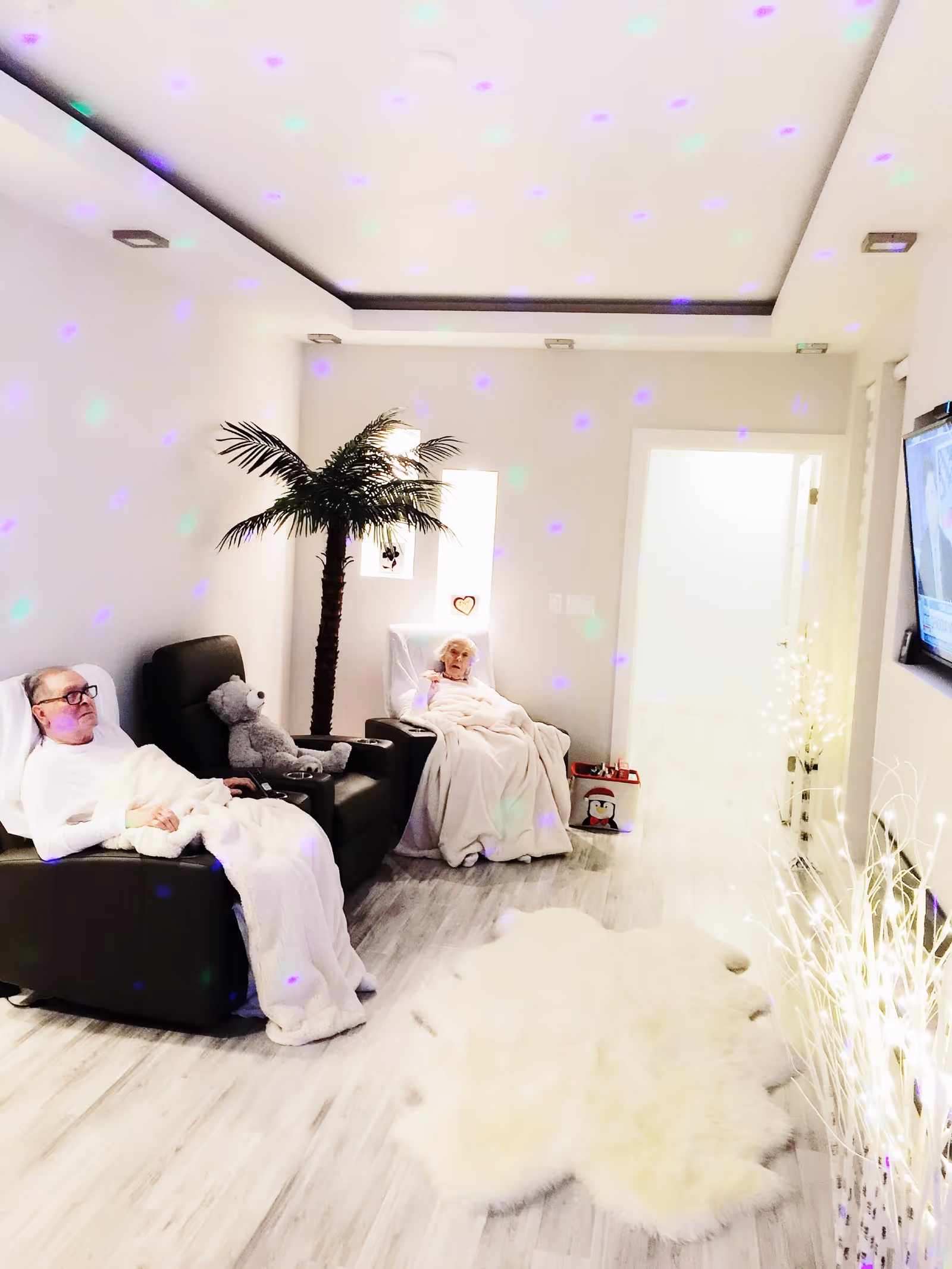 A cozy living room with two elderly people sitting in recliner chairs covered with white blankets. There is a large teddy bear on one chair, a decorative artificial palm tree, a fluffy white rug on the floor, and a TV mounted on the wall. The room has soft lighting with colorful light spots projected on the walls and ceiling.