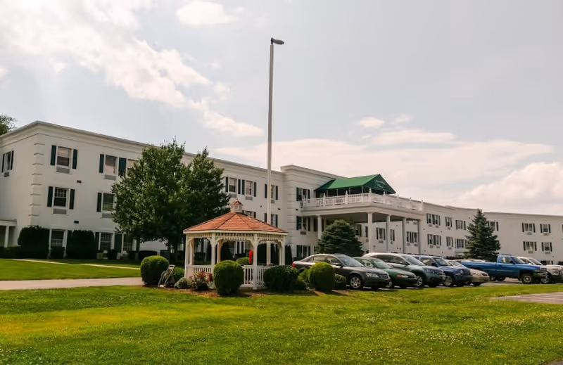 Exterior view of a large white senior living facility building with multiple windows and a green awning over a balcony. In front of the building is a small white gazebo with a red roof, surrounded by bushes and trees. Several cars are parked in a parking lot adjacent to the building, and a tall light pole stands near the gazebo. The sky is partly cloudy.