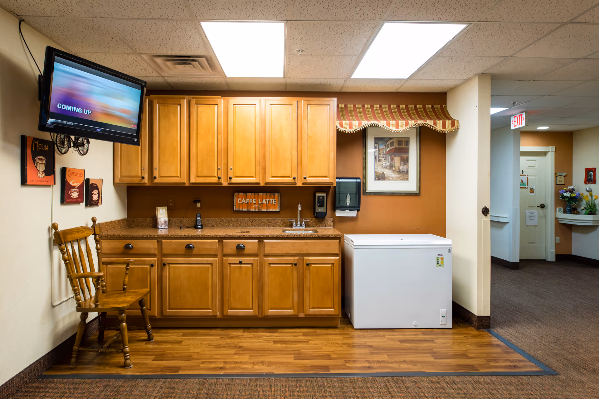 Interior view of a kitchenette area with wooden cabinets, a countertop with a sink, a white chest freezer, a wooden chair, and a wall-mounted TV. The walls have coffee-themed decorations and a framed picture. The floor is a combination of wood and carpet, and there is an exit sign visible in the hallway.