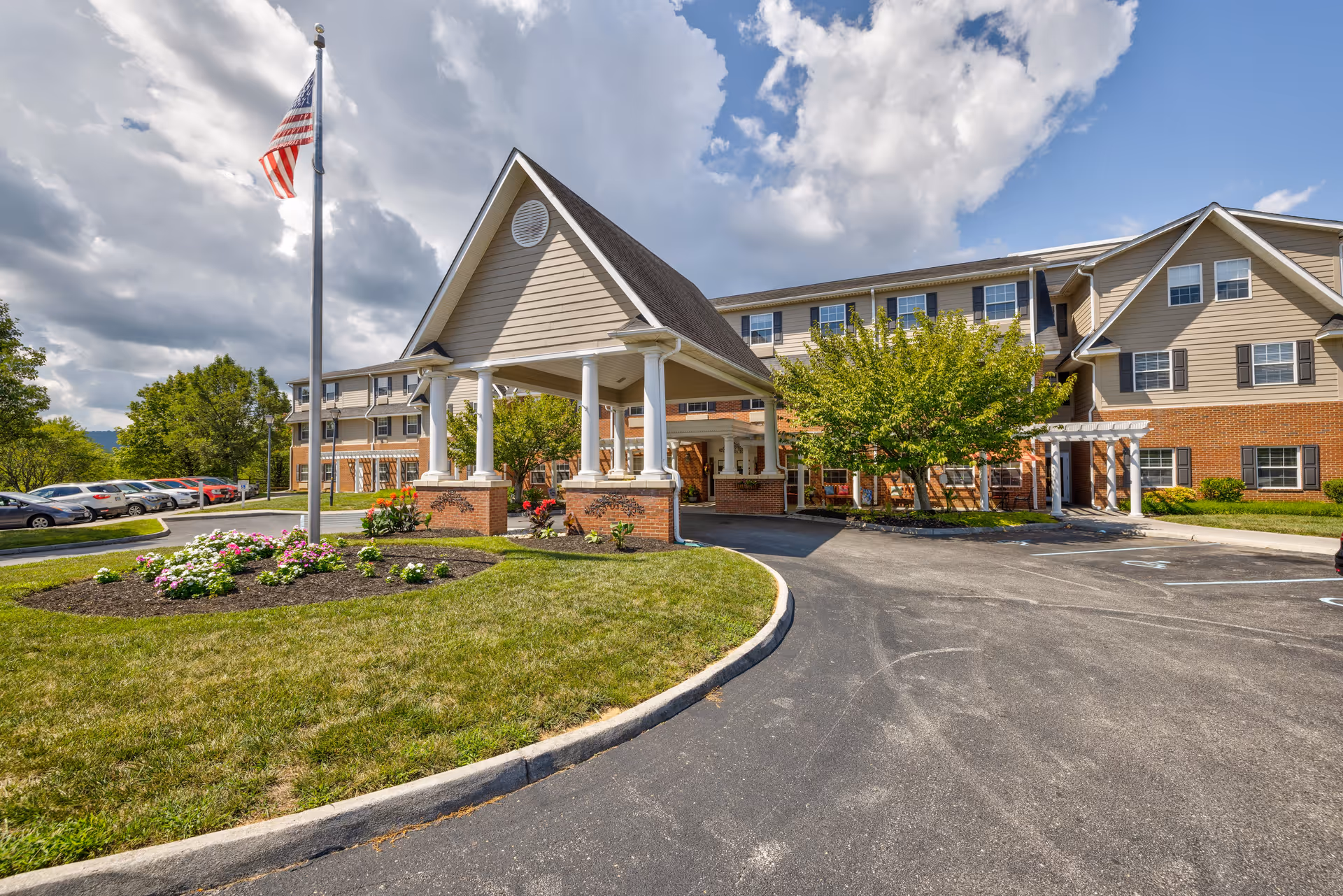 Exterior view of TerraBella Pheasant Ridge facility showing a large building with beige siding and brick accents under a partly cloudy sky. There is a covered entrance supported by white columns, a flagpole with an American flag, landscaped flower beds, a parking lot with several cars, and green trees around the building.