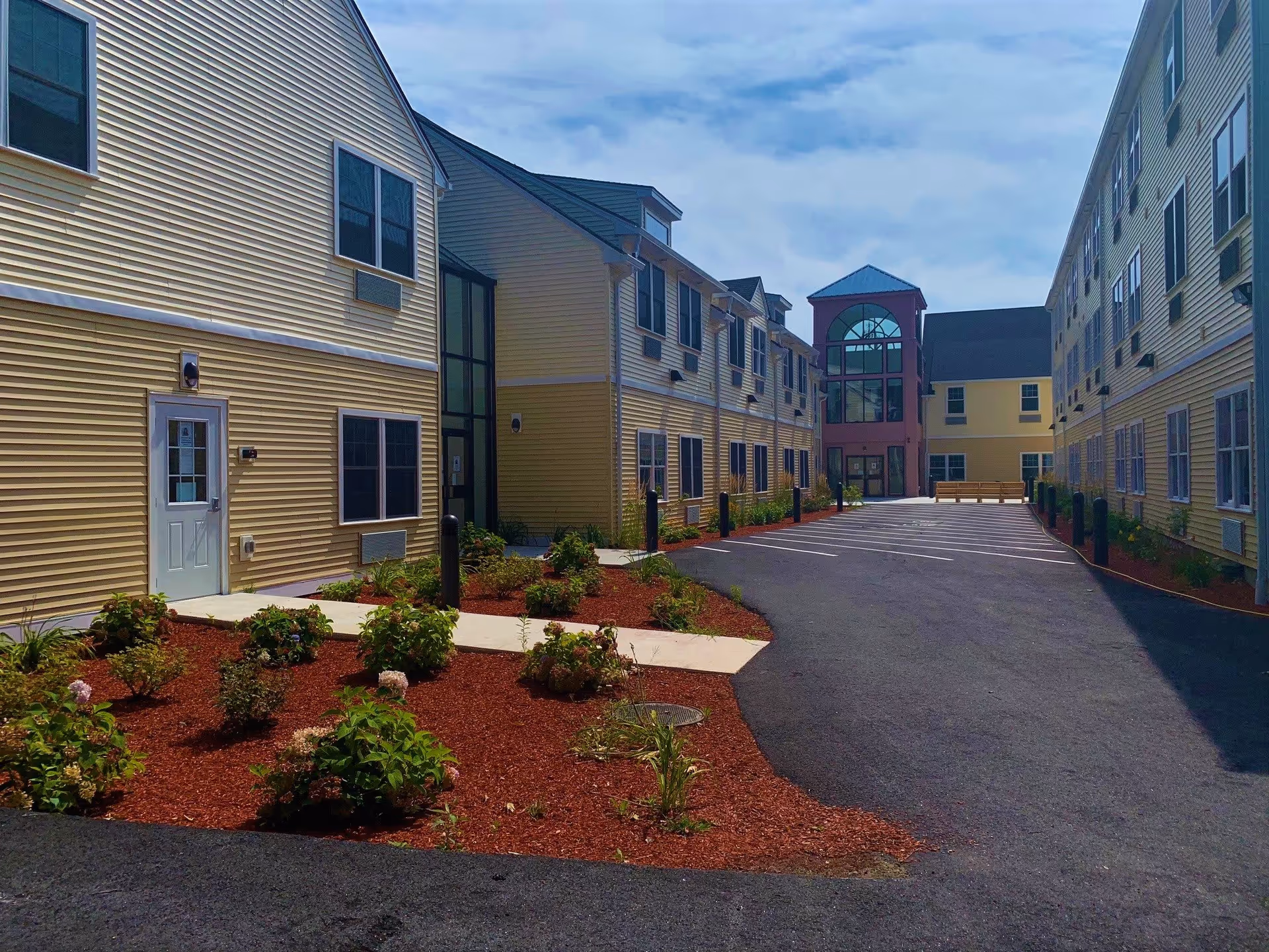 Exterior view of a senior living facility with two yellow buildings facing each other, a paved driveway in between, landscaped garden beds with shrubs and flowers, and a building entrance with large windows at the far end under a partly cloudy sky.