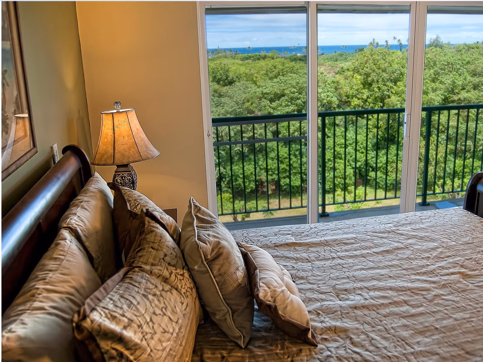 A bedroom with a neatly made bed featuring multiple pillows and a decorative bedspread. A bedside table with a lamp is next to the bed. Large sliding glass doors open to a balcony with a black railing, overlooking lush green trees and a distant view of the ocean under a blue sky.