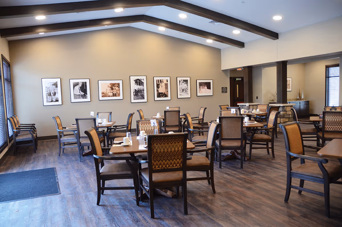 A spacious dining room with multiple wooden tables and cushioned chairs arranged neatly. The room has wooden flooring, a high ceiling with exposed beams, and several black and white framed photographs on the far wall. Large windows with blinds allow natural light to enter the room.