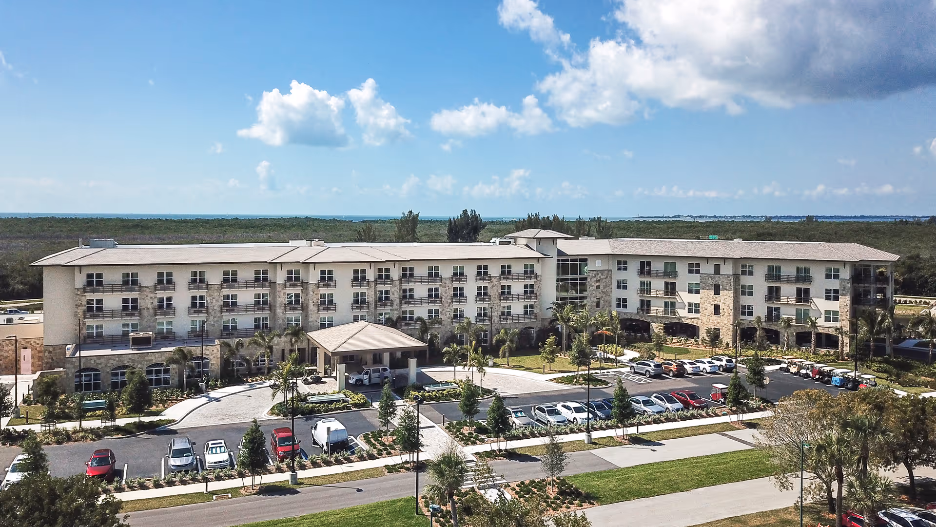 A large, modern senior living facility building with multiple floors and balconies, surrounded by landscaped greenery and a parking lot with several cars. The sky is partly cloudy with blue skies visible.