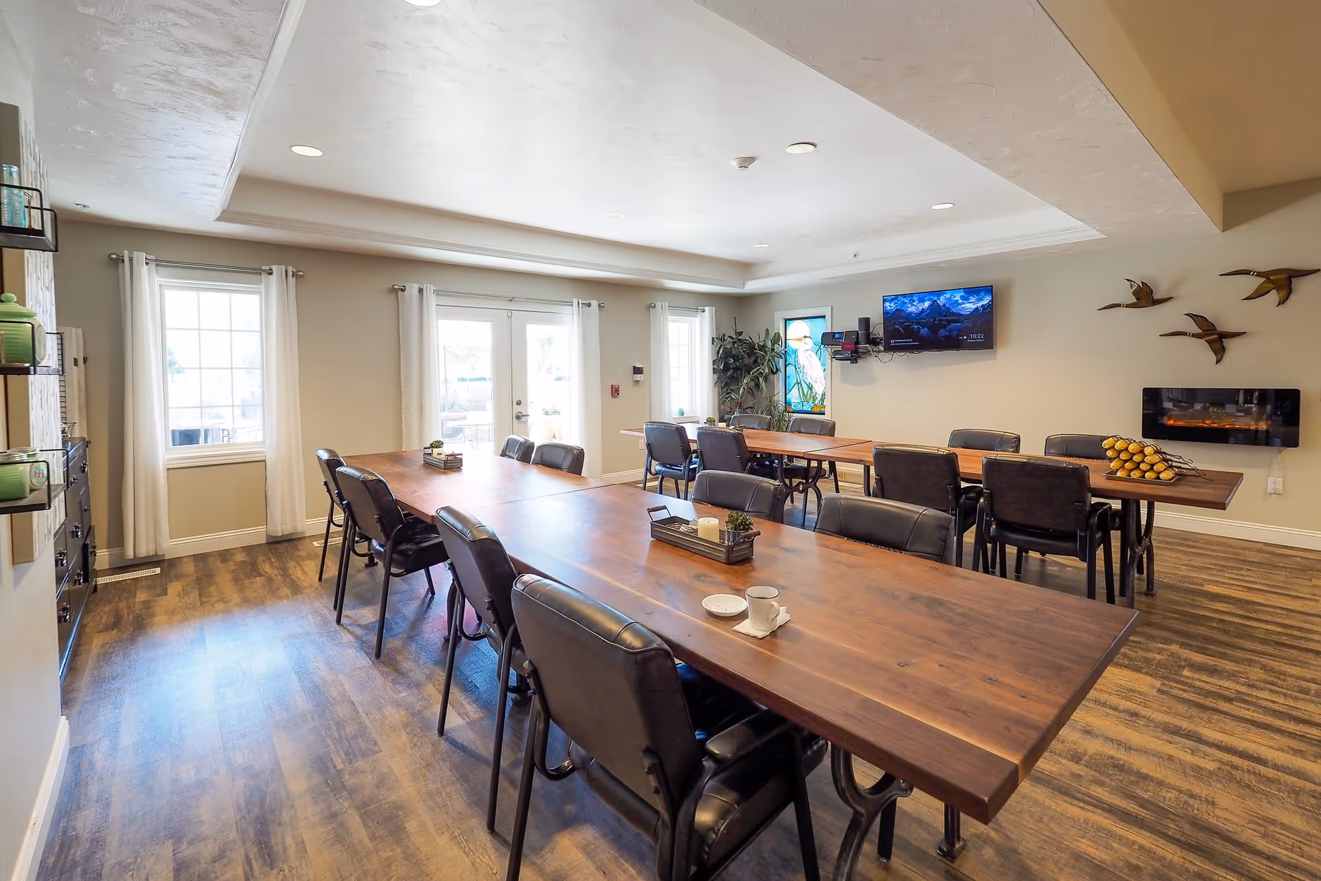 Communal dining/activity room with long wooden tables and chairs, a wall-mounted TV, and decorative wall art.