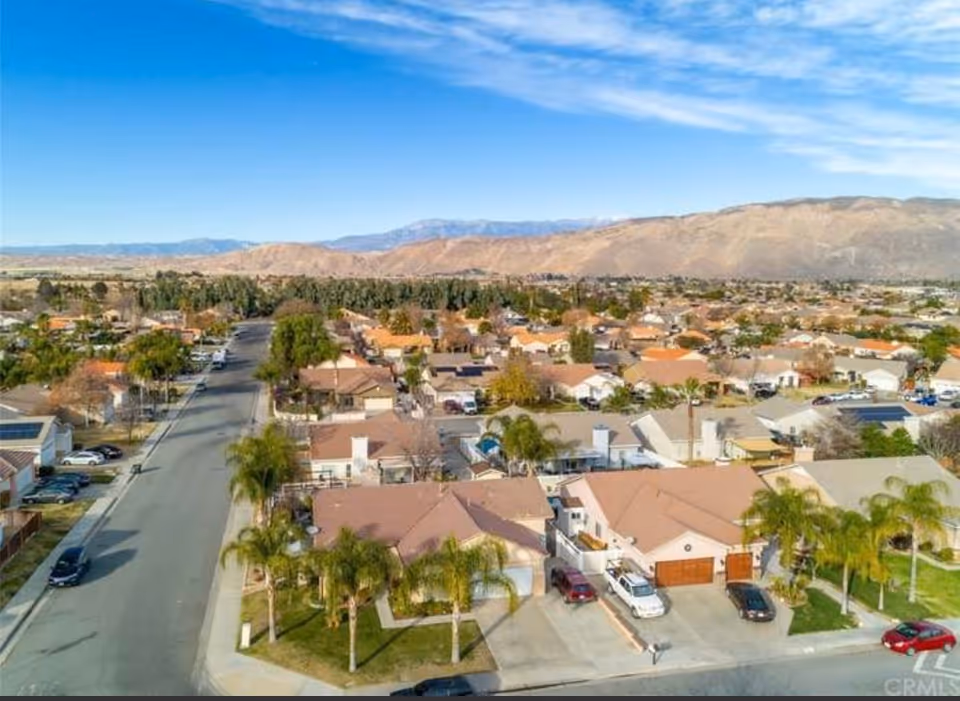 Aerial view of a suburban neighborhood with single-family homes, palm trees, and a street running through the area. In the background, there are mountains under a blue sky with some clouds.