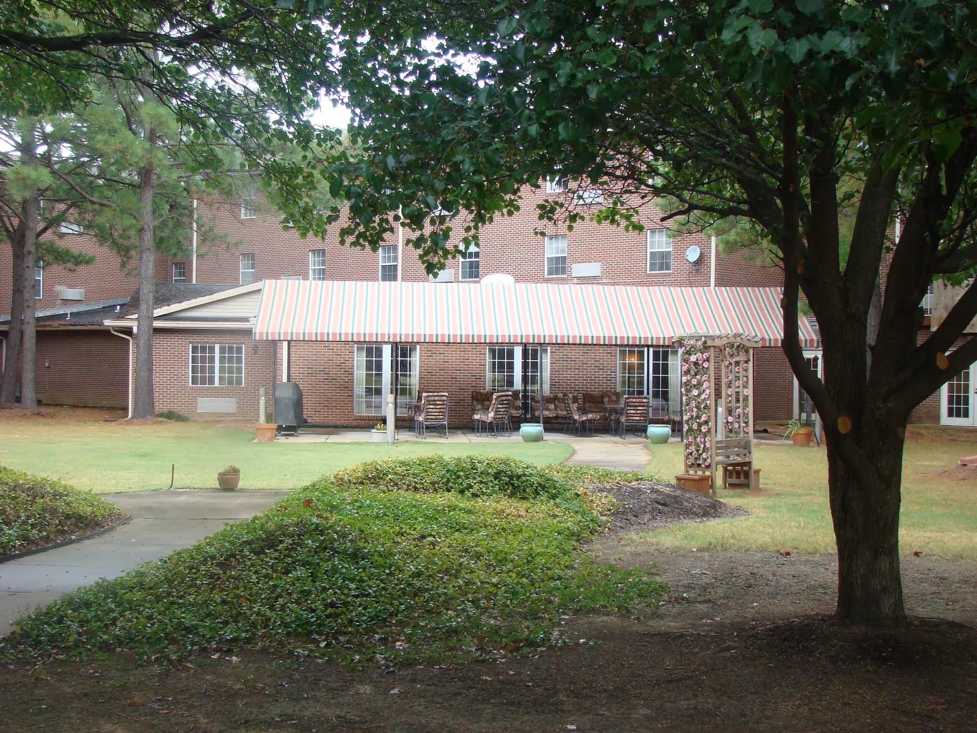 View of an outdoor patio area at Kennington Pointe with a striped awning covering several tables and chairs. The patio is surrounded by a grassy lawn, trees, and some landscaping including a trellis with flowers.