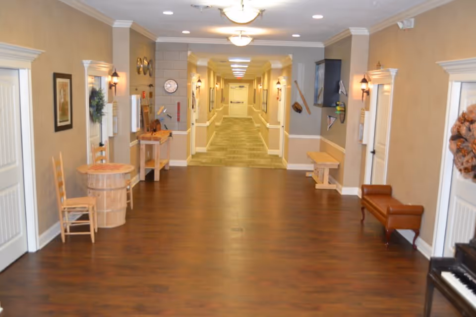 A wide hallway in a senior living facility with wooden flooring and beige walls. The hallway is decorated with framed pictures, wall sconces, and wreaths on doors. There are chairs, small tables, and a piano along the sides. The hallway leads to a carpeted corridor with more doors and lighting fixtures on the ceiling.