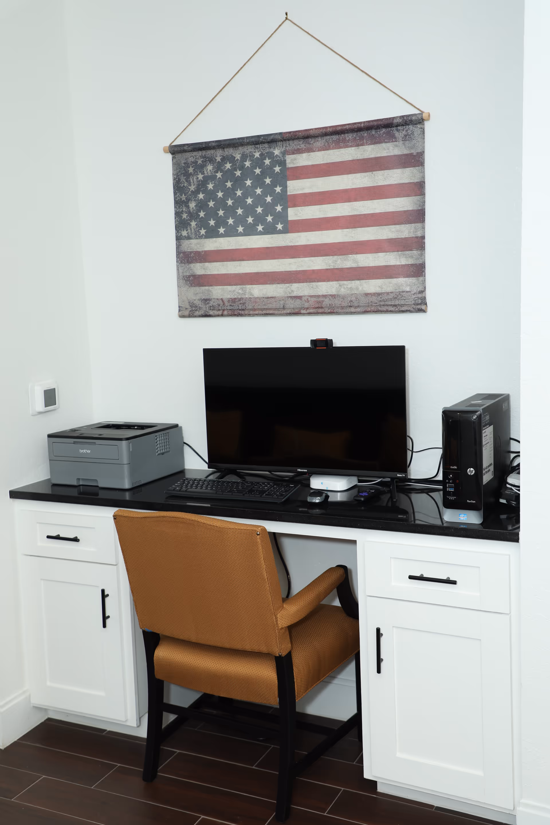 A small office workspace with a brown upholstered chair in front of a black countertop desk. On the desk are a computer monitor, keyboard, mouse, desktop computer tower, and a Brother printer. Above the desk hangs a vintage-style American flag wall decoration.