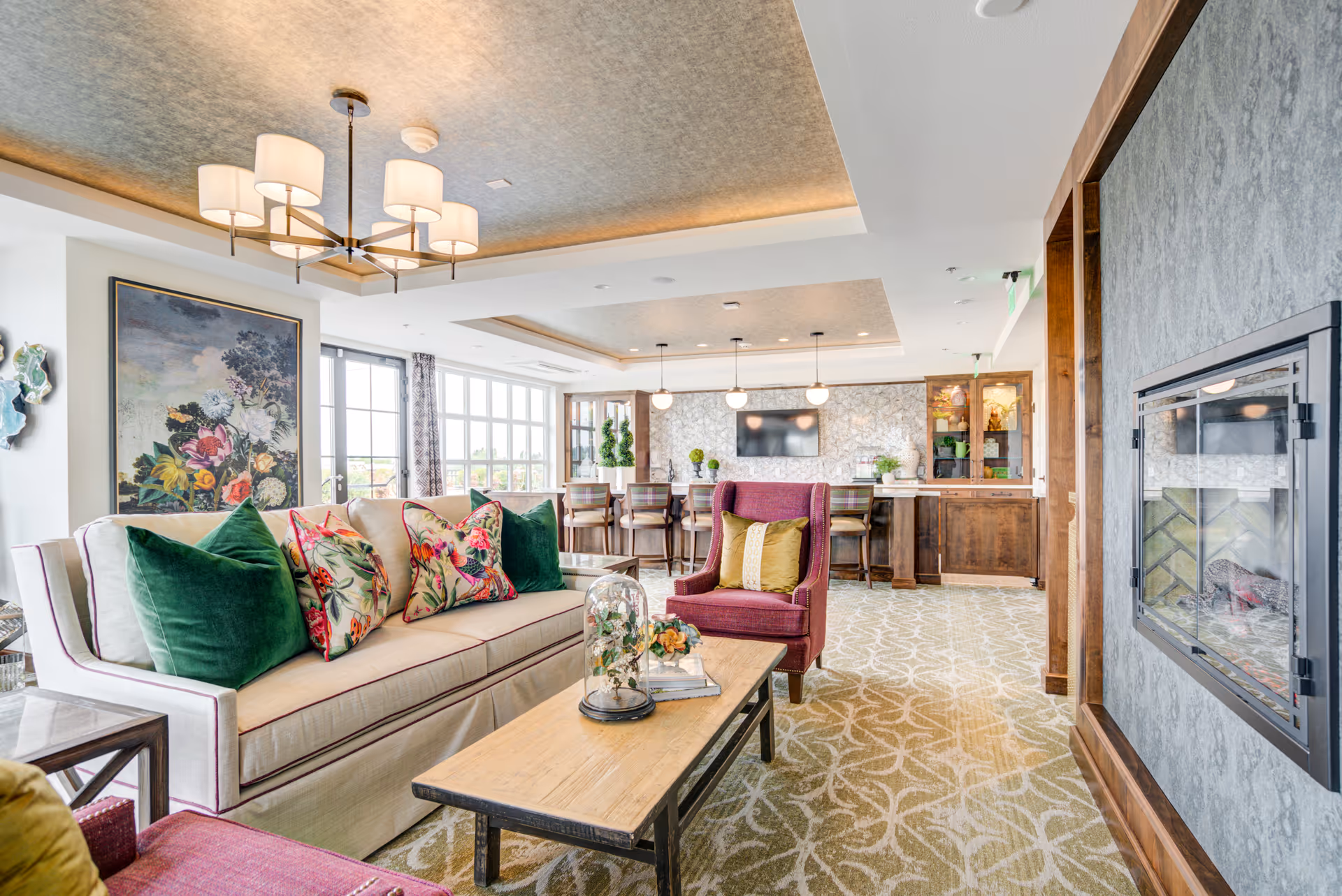 A bright and spacious living room area in a senior living facility featuring a beige sofa with green and floral pillows, a red armchair with a yellow pillow, a wooden coffee table with decorative items, a modern fireplace on the right wall, and a bar area with stools and pendant lights in the background. Large windows allow natural light to fill the room, and the ceiling has recessed lighting and a chandelier.