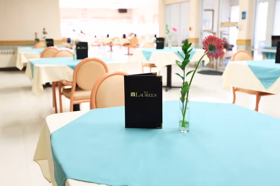 A bright dining room with tables covered in white and turquoise tablecloths. Each table has a small vase with a single flower and a black menu standing upright. The chairs are light pink with rounded backs. The room is spacious and well-lit with natural light coming from large windows.