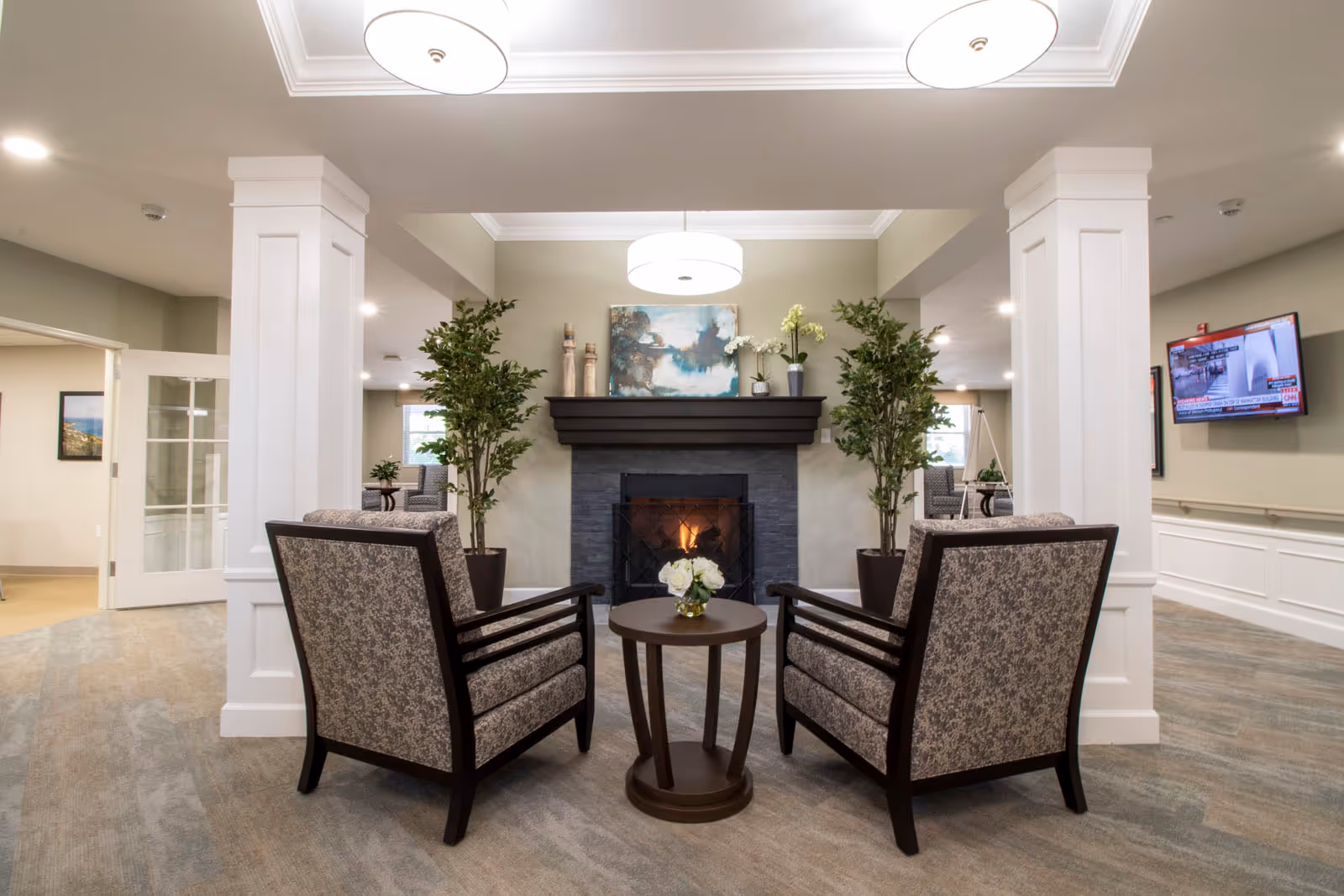 A cozy seating area in a senior living facility with two patterned armchairs facing a small round wooden table with a vase of white flowers. Behind the seating area is a lit fireplace with a dark mantel, decorated with plants and a painting above it. The room has light-colored walls, carpeted floors, and ceiling lights. A TV is mounted on the wall to the right, and there are large white columns framing the seating area.